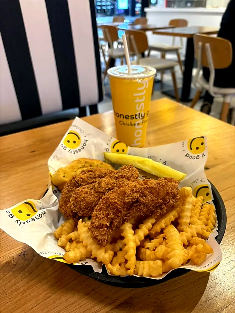 An Honestly Good box: hand-breaded chicken fingers, crinkle-cut fries, a pickle spear and Texas toast garlic bread, with a yellow Honestly Good cup behind it
