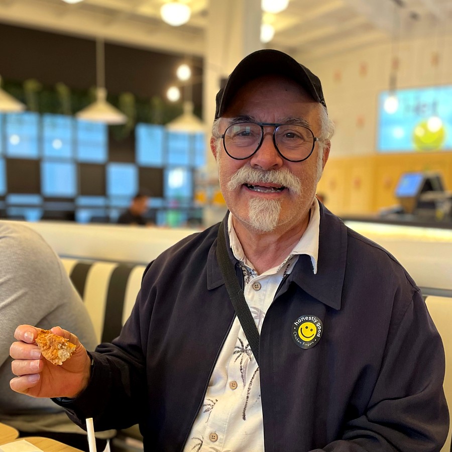 A guest wearing an Honestly Good badge pin, smiling in the dining room with a chicken finger in his hand